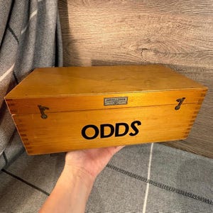Puede incluir: Una caja de madera con la palabra "ODDS" en letras negras. La caja tiene cierres metálicos y una pequeña etiqueta rectangular. La caja está sostenida por una persona.