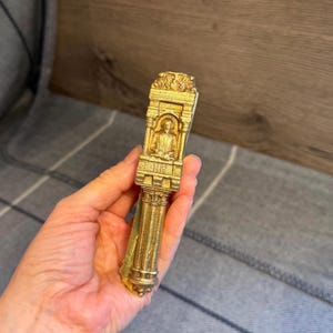 May include: A gold-toned metal object with a vertical, column-like structure featuring an architectural archway. Inside the arch is a relief figure of a person, with the numbers 1564 and 1616 engraved below the figure. The piece has a textured finish.