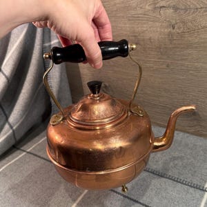 May include: A copper tea kettle with a black wooden handle and a matching black knob on the lid. The kettle has a curved spout and sits on three small brass feet. The handle is held by a hand.