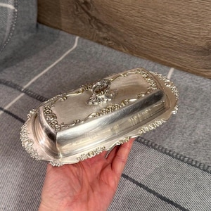 May include: Silver-plated butter dish with a decorative floral design. The rectangular dish has a lid with a handle and ornate detailing around the edges. The butter dish is held in a hand.