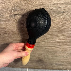 Pode incluir: Uma maraca preta com uma superfície texturizada e um cabo de madeira. A maraca tem uma faixa vermelha perto do cabo e é projetada para uso musical. O instrumento é segurado em uma mão.