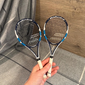 May include: Two Babolat tennis rackets with navy blue frames, white accents, and blue detailing. The rackets have black stringing and white grips. The rackets are held against a gray and white striped blanket.