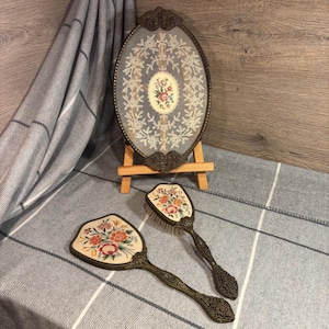 Puede incluir: Un juego de tocador antiguo con un espejo ovalado enmarcado y dos cepillos a juego. El espejo y los cepillos tienen marcos de metal ornamentados y diseños florales bordados en tonos de rosa, naranja y verde. El conjunto se exhibe sobre una tela a rayas grises y blancas.