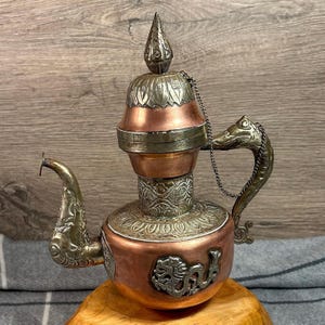 Puede incluir: Una tetera de cobre y plata con intrincados diseños florales y de dragones. La tetera tiene un pico en forma de cabeza de dragón y una tapa con un pomo.