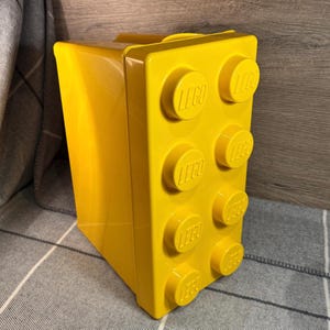 May include: A bright yellow plastic storage box shaped like a LEGO brick. The box has six LEGO studs on the front, each with the LEGO logo printed on it.