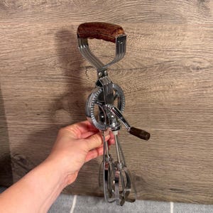 Könnte beinhalten: Ein manueller Vintage-Schneebesen mit Kurbel, bestehend aus einem polierten Chrom-Metallrahmen, einem dunkelbraunen Holzgriff an der Oberseite und einem passenden dunkelbraunen Holzkurbelknopf. Das Küchengerät hat zwei Zahnräder und zwei Drahtbesen.