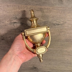 Op de afbeelding: Een goudkleurige metalen deurklopper met een centraal urnvormig lichaam, een rechthoekig naamplaatje met een touwachtige rand en een ronde slagplaat aan de onderkant. Het ontwerp bevat een decoratieve knop aan de bovenkant en een gebogen handvat.