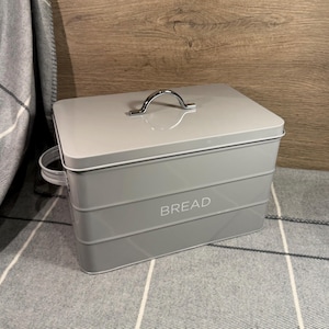 Puede incluir: Una panera rectangular de metal gris claro con tapa extraíble que presenta un asa arqueada de tono plateado. La parte frontal del recipiente tiene grabada la palabra BREAD en letras mayúsculas blancas e incluye un asa lateral para transporte.