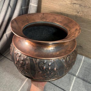 May include: A large, copper planter with an ornate, embossed design. The planter has a wide, flared rim and a rounded body. The copper has a warm, aged patina.