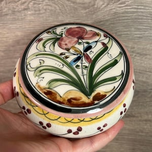 May include: A round ceramic trinket box with a lid. The lid features a floral design with pink, green, and blue accents on a white background. The box has a pink and yellow band with maroon dots around the base.