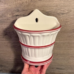 May include: A white ceramic wall pocket with a red trim. The pocket has a fluted design and a small hole at the top for hanging. The pocket is held by a hand against a wooden background.