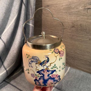 Könnte beinhalten: Ein cremefarbenes Keramikgefäß mit silbernem Deckel und Griff. Das Gefäß hat ein gemaltes Pfauenmuster in Blau, Lila und Rosa. Der Griff hat ein geschwungenes, herzförmiges Design.