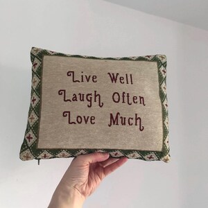 May include: A rectangular cushion featuring the embroidered words "Live Well Laugh Often Love Much" in burgundy. The cushion has a beige centre and a green border with red and beige diamond patterns.