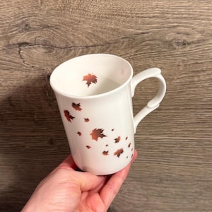 Könnte beinhalten: Weiße Keramiktasse mit gebogenem Henkel, verziert mit kupferfarbenen fallenden Blättern. Die Tasse hat eine glatte, glänzende Oberfläche und ist für Heißgetränke geeignet. Ein saisonales Produkt zum Thema Herbst.