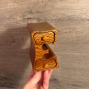 May include: A small wooden storage unit shaped like the letter E, featuring a light brown wood grain finish. The unit includes two drawers with small round knobs, one positioned at the top and one at the bottom, with an open space in the middle.