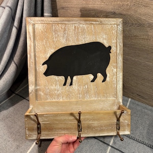 Puede incluir: Un organizador de pared rústico de madera con un acabado envejecido. Presenta una silueta negra de un cerdo en el panel superior y tres ganchos de color bronce debajo para colgar objetos. El diseño tiene una estética de granja.