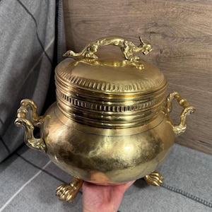Può includere: Un contenitore vintage in ottone con coperchio e due manici. Il coperchio è sormontato da una figura decorativa di animale. Il contenitore ha tre piedi ad artiglio. L'ottone ha una finitura lucida.