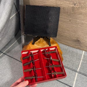 May include: A red box containing a set of small, vintage appetizer forks and knives with wooden handles and silver blades. A small black chalkboard on a wooden easel is in the background.