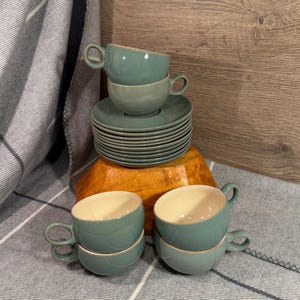 May include: A set of sage green ceramic cups and saucers. The cups feature a rounded body with a circular handle and a cream-colored interior. A stack of matching saucers sits beneath one cup, while two pairs of cups are arranged in the foreground.