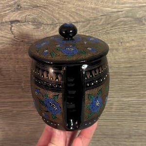 Op de afbeelding: Een decoratieve pot in zwart en bruin, versierd met blauwe bloempatronen. De pot heeft een deksel en een afgeronde vorm. Het bloemmotief herhaalt zich op het deksel en de body van de pot. De pot wordt in een hand gehouden.