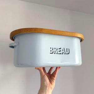 Può includere: Un portapane azzurro con coperchio in legno. La parola "BREAD" è stampata in nero sul davanti. Il portapane ha una forma rettangolare con angoli arrotondati e piccole maniglie. Il coperchio è in legno chiaro.