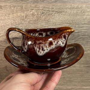 Könnte beinhalten: Eine braune Keramik-Sauciere mit passender Untertasse. Die Sauciere hat einen Griff und einen Ausguss. Der Rand der Sauciere und der Untertasse hat ein weißes und braunes Sprenkelmuster. Das Set steht auf einer Holzoberfläche.