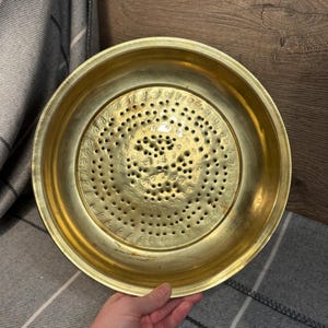 May include: A round, brass strainer with a decorative pattern of holes and numbers. The strainer is approximately 25 cm in diameter and has a slightly raised rim.
