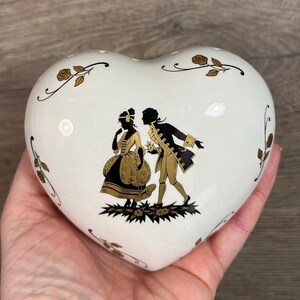 May include: White ceramic heart-shaped trinket box with a gold and black silhouette of a couple in 18th-century attire. The box is decorated with gold floral accents.