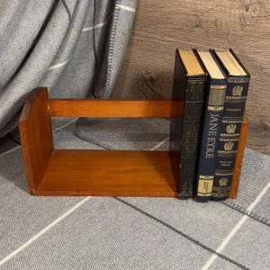 Könnte beinhalten: Ein kleiner, brauner Holz-Bücherregal mit drei antiken Büchern. Das Bücherregal hat ein einfaches Design. Die Bücher sind dunkel mit goldener Schrift, darunter Titel wie "Jane Eyre" und "Krieg und Frieden".