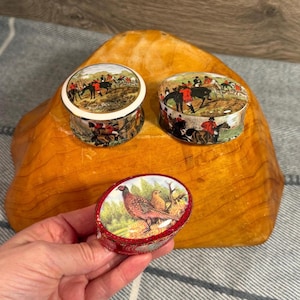 May include: Three small, round, and oval lidded trinket boxes featuring detailed hunting scenes. Two boxes display riders on horseback in red coats, while the third oval box features a colourful pheasant bird in a natural outdoor setting.