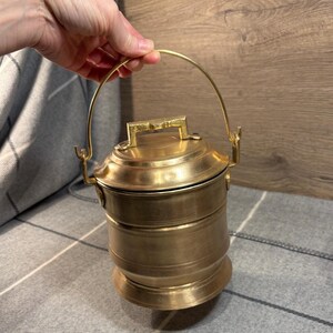 May include: A vintage brass container with a handle. The container has a cylindrical shape with a lid and a base. The handle is arched and attached to the top of the container. The brass has a warm, golden color.