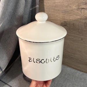 Op de afbeelding: Een witte keramische mok met een zwart handvat en de tekst "But First, Coffee" in een zwarte, vette letter. De mok staat op een wit oppervlak. De mok is ongeveer 10 cm hoog.