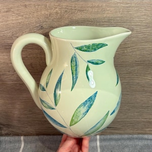 May include: A light green ceramic pitcher with a large handle and a spout. The pitcher is decorated with blue and green leaf designs. The pitcher is held by a person.