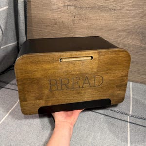 May include: A rectangular bread bin featuring a matte black metal body and a natural wood front panel. The word BREAD is engraved in a simple font on the centre of the wooden panel, which also includes a horizontal slot handle at the top.