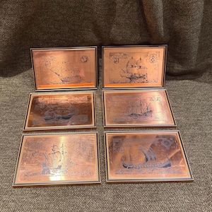 May include: Six framed copper plaques with ship illustrations. The plaques feature various sailing vessels, including the "Victory" and "Santa Maria." Each plaque has a dark border and a copper-colored interior, creating a vintage aesthetic.