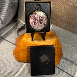 May include: A round, gold-framed picture of a street scene with buildings and people. The picture is in a black box with a gold stand.