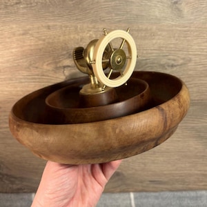 May include: A wooden nutcracker with a brass ship's wheel and a brown wooden bowl. The nutcracker is in the shape of a ship's wheel and has a brass handle. The bowl is made of wood and has a dark brown finish.