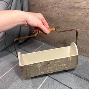 May include: A rectangular, vintage-style wooden box with a metal handle and a wooden grip. The box is painted in a distressed gray and cream color scheme, with French text and floral accents. The handle is made of dark metal.