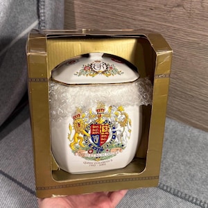 May include: A white ceramic jar with a lid, featuring a royal crest and the text "Queen Elizabeth II 1952-2002." The jar is in a gold box, with bubble wrap inside. The lid has floral designs and the initials "ER".
