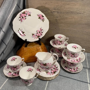 Può includere: Un servizio da tè in porcellana con base bianca, motivi a rose rosa e bordi argentati. Il set comprende un grande piatto da portata, diverse tazze da tè con piattini abbinati e una lattiera, decorati con delicati fiori rosa.