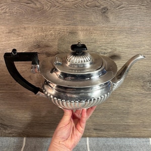 May include: A silver teapot with a black handle and lid knob. The teapot has a rounded body with a decorative beaded edge. The spout is curved, and the lid has a small, ornate finial. The teapot is held by a person.
