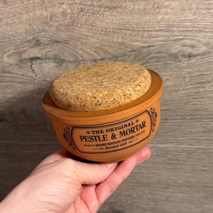 May include: A terracotta pestle and mortar set with a cork lid. The mortar has a rounded shape and features the text "THE ORIGINAL PESTLE & MORTAR" in a decorative banner. The set is held in a hand, showcasing its size and design.