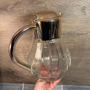 May include: A clear glass water jug with a silver-coloured metal handle, lid, and rim. The jug has a bulbous shape with a textured, wavy design. The lid has a small spout. The jug is held by a person.