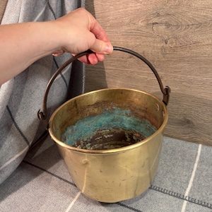 Puede incluir: Un cubo de latón vintage con un asa de metal oscuro. El interior muestra signos de envejecimiento con oxidación azul-verdosa. El cubo está sostenido por una mano, lo que sugiere su tamaño y portabilidad. La estética general es rústica y antigua.