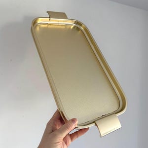 May include: A rectangular gold-colored serving tray with rounded corners and a textured surface. The tray has two handles on the short sides. The tray is held by a person's hand.