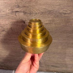 May include: A set of six circular brass weights stacked in a pyramid formation, with the largest weight at the base and the smallest at the top. Each individual weight has a smooth, metallic gold-toned finish and a flat, cylindrical shape.