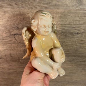 May include: A ceramic angel figurine with wings, sitting with one leg crossed over the other. The angel is painted in shades of cream, yellow, and gold.
