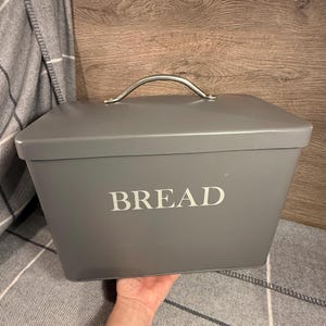 May include: A grey rectangular bread bin with a silver handle. The word "BREAD" is printed in white on the front. The bread bin is on a grey and white plaid blanket. The bread bin is made of metal.