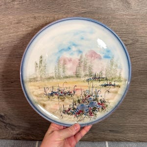 Puede incluir: Un plato redondo de cerámica con una escena de paisaje pintada a mano. El plato presenta un borde azul claro y un diseño central de un campo con árboles, montañas y flores coloridas. La paleta de colores incluye azul, rosa, verde y amarillo.