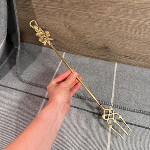 以下が含まれることがあります： 小さなフィギュアの形をした装飾的なハンドルが付いた、長く細い真鍮製の暖炉用フォーク。この道具は、真っ直ぐで細いシャフトと、タインの付け根に複雑な透かし彫りの幾何学模様が施された3本爪のヘッドを備えています。